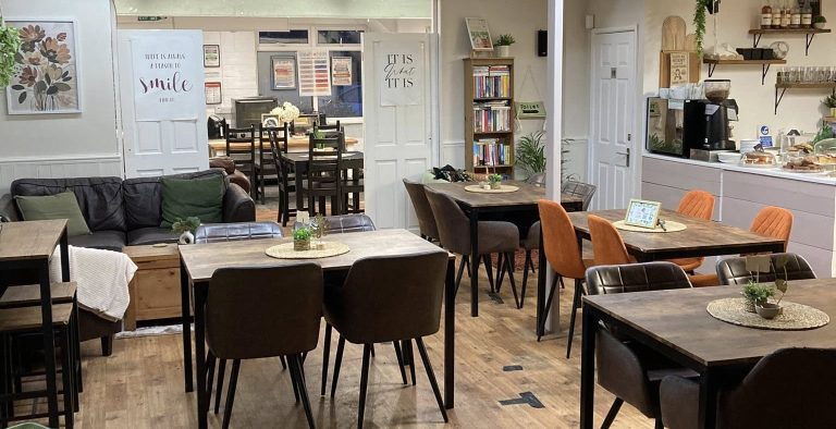 Interior of The Quirky Cafe in Hoylake, showing a warm, inclusive community space with wooden tables, mixed seating, plants, bookshelves and a relaxed café layout.